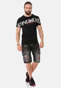 Schwarzes T-Shirt mit kurzen Ärmeln und weißem Grafiktext, kombiniert mit grauen Cargo-Shorts mit Reißverschlüssen und roten Akzenten. Schwarze Sneakers mit pinken Details.