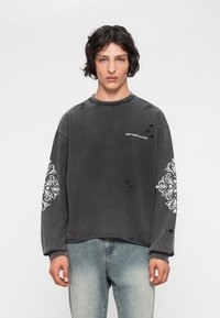 Joven con cabello rizado, lleva un sudadera negra desgastada con patrones blancos en las mangas y el texto "NO DES PERDER CULTURA", combinada con jeans desgastados.