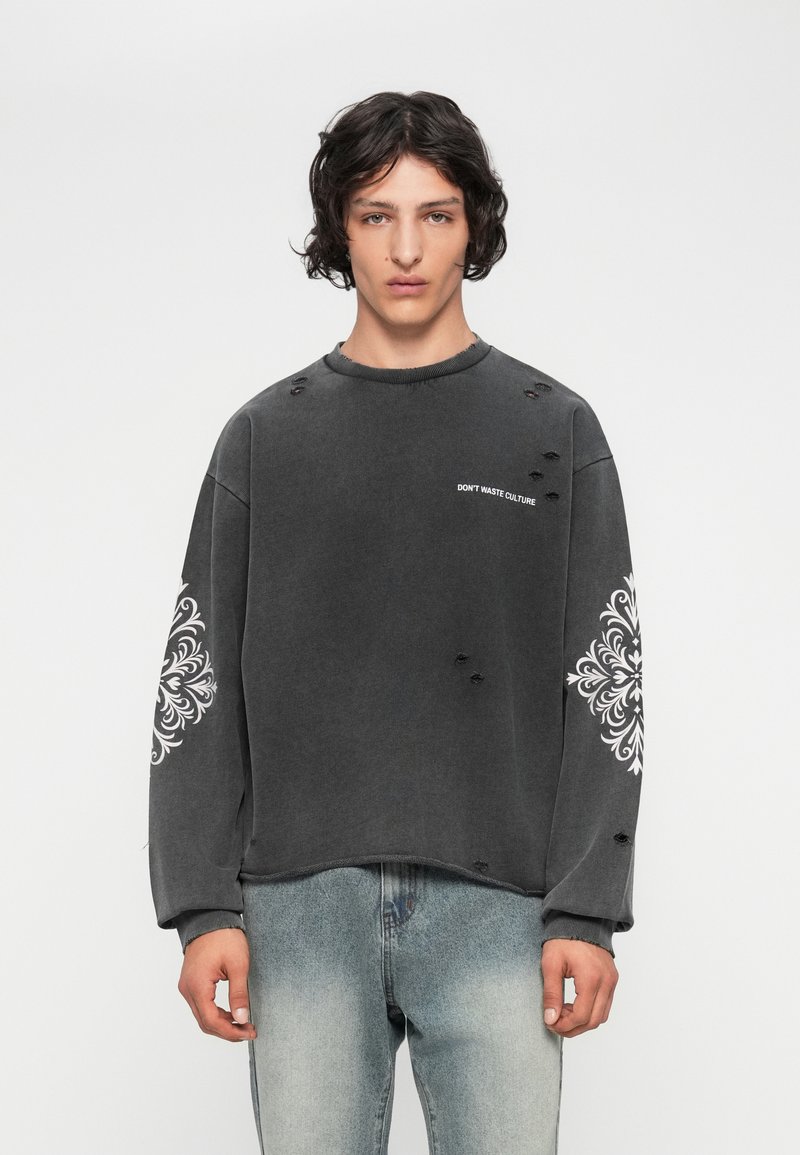 Joven con cabello rizado, lleva un sudadera negra desgastada con patrones blancos en las mangas y el texto "NO DES PERDER CULTURA", combinada con jeans desgastados.