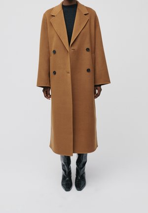 Manteau couleur chameau, à double boutonnage, avec de grands revers et des boutons noirs, présentant une coupe décontractée et atteignant le mi-mollet, associé à des bottes noires.
