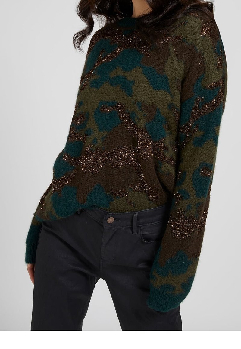 Femme portant un pull à motif camouflage duveteux aux couleurs vert foncé, marron et sarcelle, associé à un pantalon foncé, sur un fond uni.