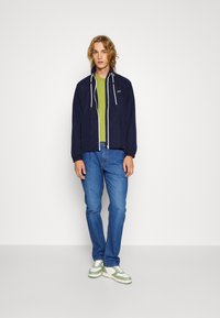 Veste coupe-vent marine avec des cordons blancs, superposée à un t-shirt vert. Portée avec un jean bleu et des baskets avec des accents verts. Fond neutre.