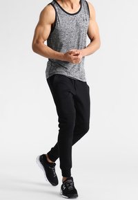 Hombre con camiseta atlética sin mangas gris, pantalones de chándal negros y zapatillas de running negras caminando frente a un fondo blanco.