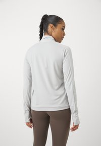 Lys grå langærmet atletisk top med høj krave, blød tekstur og Nike-logo på ryggen. Kombineret med brune leggings.