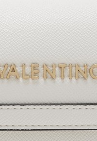 Close-up van roomkleurig gelooid leer met gestikte details en gouden metalen letters die "VALENTINO" spellen.