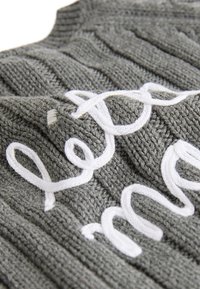 Pull gris en maille avec texture côtelée en relief, orné de broderies blanches formant un texte sur le devant. Détail en gros plan visible.