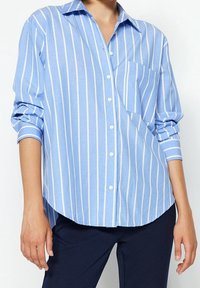 Ljusblå skjorta med vita vertikala ränder, som har en ficka på bröstet och uppvikta ärmar. Mjuk tyg, avslappnad passform, rundad nederdel.