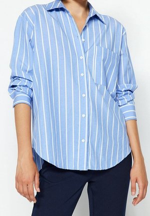 Camisa de botones azul claro con rayas verticales blancas, que presenta un bolsillo parche y mangas enrolladas. Tela suave, corte relajado y dobladillo curvado.