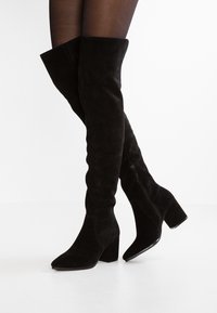 Mustat mokkanahkaiset yli polven saappaat, joissa on terävä kärki ja tuellinen korko. Saappaissa on sivuvetoketju ja pehmeä, tekstuurinen pinta.