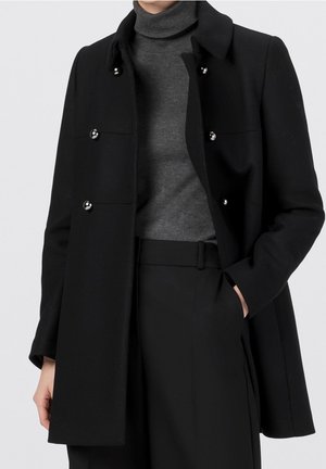 Personne portant un manteau noir croisé avec des boutons argentés, un pull à col roulé gris, et un pantalon noir taille haute, main dans la poche.