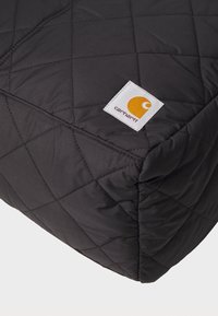 Coussin noir matelassé avec un léger éclat, arborant une étiquette blanche avec un logo orange. Texture lisse avec un motif de quilting en forme de diamant.