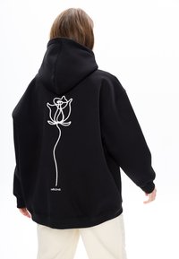 Zwarte hoodie met een grote witte rozenomtrek op de achterkant. Voorzien van een kangoeroezak en ribgebreide manchetten. Zacht, textuurachtig materiaal.