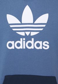 Blue sweatshirt with white Adidas logo featuring a three-leaf insignia and horizontal stripes. Material: soft fabric, textured finish.