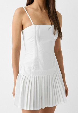 Woman wearing a white sleeveless dress with thin straps, a fitted pleated bodice, and a pleated skirt.