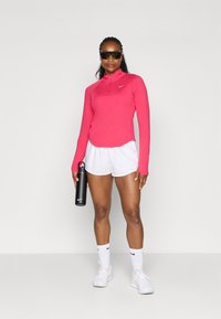 Pink duguljasti sportski top s patentnim zatvaračem; uparen s bijelim šortsem, bijelim čarapama i svijetlim tenisicama, drži crnu bocu za vodu.