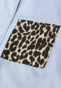 Chemise bleu clair avec une poche carrée cousue ornée d'un motif léopard noir et marron sur fond beige.