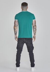 T-shirt fitted color verde acqua con rifiniture bianche, abbinato a pantaloni cargo neri. L'outfit presenta una texture liscia e un design moderno.