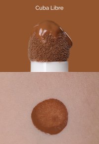 Base líquida en un aplicador redondeado con una punta suave y texturizada, mostrando un tono marrón intenso sobre un fondo de tono de piel.