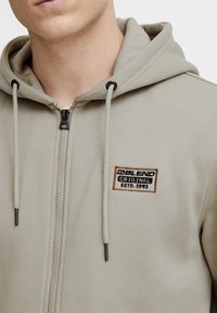Sudadera con cremallera de color gris claro y textura suave, que presenta un parche de logo bordado en marrón oscuro y negro. Capucha ajustable con cordones.