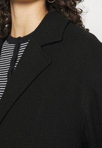 Mujer vistiendo un abrigo negro sobre una camiseta de rayas en blanco y negro, con cabello rizado y un pendiente de aro plateado visible.