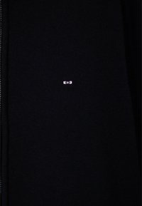 Tissu tricoté noir avec un petit logo rose brodé, centré, représentant deux yeux stylisés.