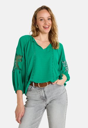 Blusa verde con escote en V, tejido texturizado, acentos de encaje en las mangas y mangas abullonadas, combinada con jeans gris claro y un cinturón marrón.