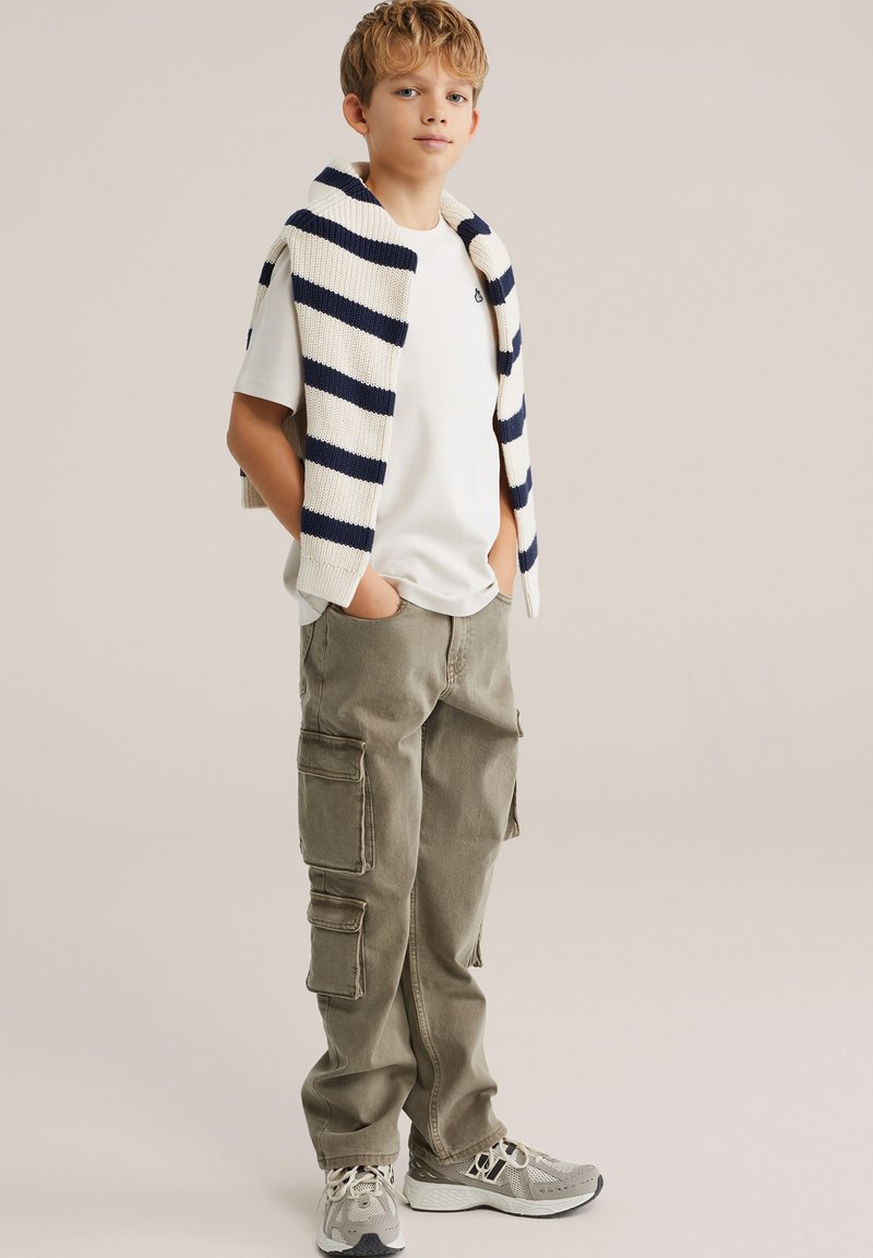 Bijela majica s okruglim izrezom, kremasto-plavi prugasti džemper bez rukava, maslinaste cargo hlače s džepovima i sive tenisice.