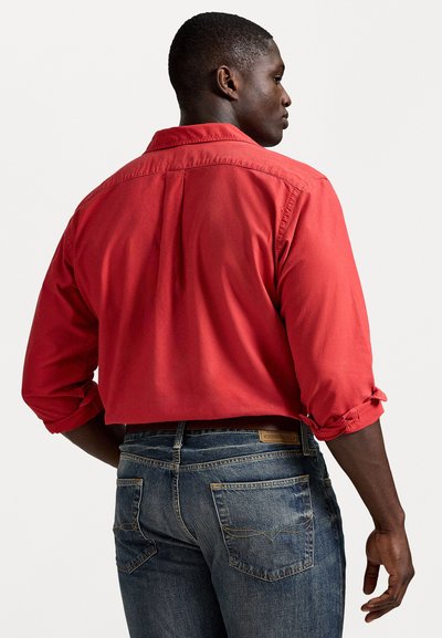 Camisa vermelha de manga longa com punhos dobrados, vista de trás. Usada com jeans azul-escuro de ganga com bolsos traseiros e detalhes de costura.