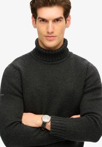 Suéter de cuello alto de canalé en gris oscuro, hecho de una tela de punto texturizada, con mangas largas y combinado con un reloj plateado en la muñeca izquierda.