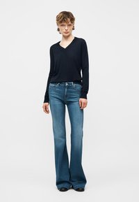 Personne portant des lunettes transparentes, un haut noir à manches longues avec un col en V, et un jean bleu taille haute évasé, debout devant un fond uni.