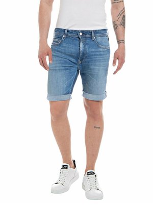 Hombre que lleva pantalones cortos de mezclilla azul claro remangados, zapatillas blancas y una camiseta blanca, con tatuajes visibles en brazos y piernas.