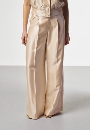 Frau trägt beige Satinhose mit hohem Bund und weitem Bein, vorne mit Falten, dazu passendes ärmelloses, knöpfbares Oberteil, steht auf grauem Boden.