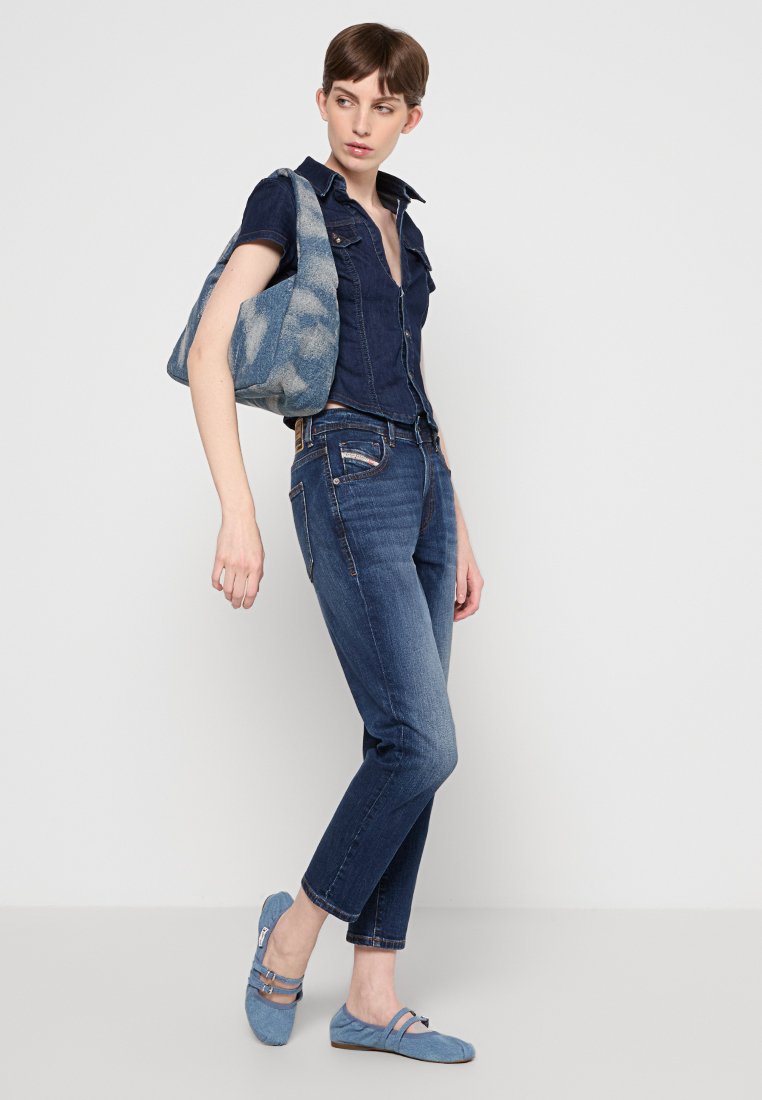 Donna con camicia in denim scuro e jeans, che porta una borsa a spalla blu con motivo e indossa ballerine azzurro chiaro.