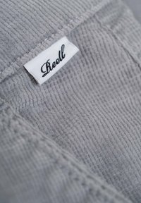 Gray corduroy fabric featuring a white tag with "Reell" in script. Texture is ribbed, emphasizing a soft yet durable material.