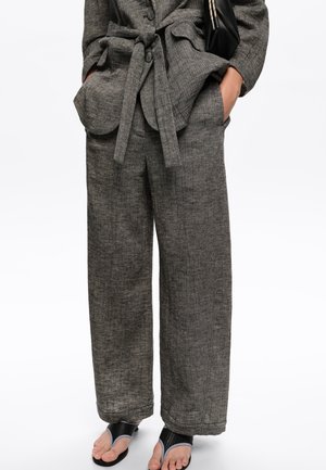 Mujer con pantalones anchos de pierna ancha y textura gris, y chaqueta a juego con cinturón, manos en los bolsillos, sandalias negras abiertas visibles.