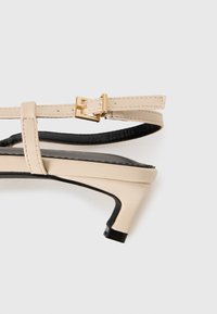 Beige høyhælte sandaler med et elegant design, svarte detaljer, smale stropper og en gullfarget spenne. Glatt skinnstruktur og spiss tå.