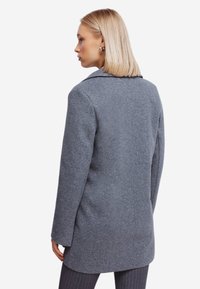 Grå uljakke med en skræddersyet pasform, indsnit krave, lange ærmer og en glat tekstur. Har en mellemlang længde og rene linjer.