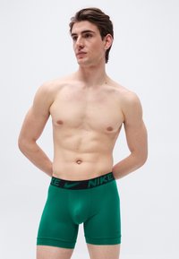 Jongetje met kort bruin haar, gekleed in groene Nike boxer shorts, staand met handen achter de rug tegen een effen witte achtergrond.