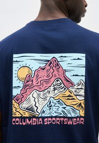 Άτομο που φοράει μπλε πουκάμισο με πολύχρωμο γραφικό βουνού, ήλιο και το κείμενο «Columbia Sportswear» στην πλάτη.