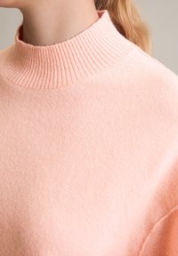 Maglione rosa tenue con un collo alto a costine, realizzato in materiale morbido. La vista ravvicinata mostra la trama e i dettagli delle cuciture sulla spalla.
