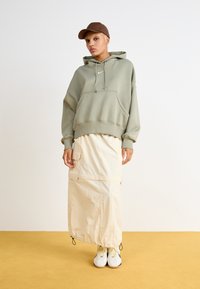 Sweatshirt oversized de cor verde-claro com capuz, bolso frontal; saia creme com fechos e bolsos laterais; sapatos brancos com atacadores amarelos.