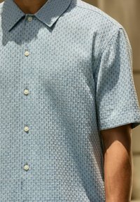 Hombre con camisa de manga corta azul claro con botones y patrón geométrico texturizado en un entorno interior bien iluminado.