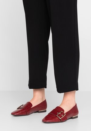 Voeten met rode loafers met krokodillenstructuur en gouden gesp, gecombineerd met zwarte cropped broeken tegen een effen achtergrond.