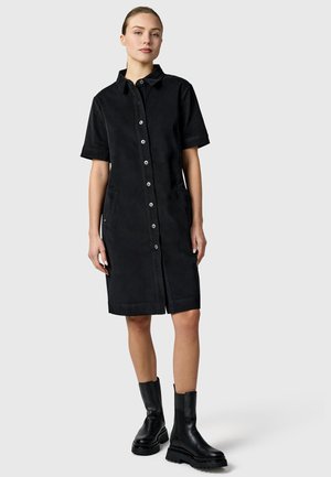 Kobieta stojąca, ubrana w czarną sukienkę na guziki do kolan z krótkimi rękawami oraz czarne masywne botki za kostkę na jednolitym tle.