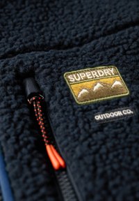 Gros plan sur une veste polaire bleu marine texturée avec un patch de logo montagne "Superdry" brodé et une fermeture éclair noire avec des cordons de tirage orange.