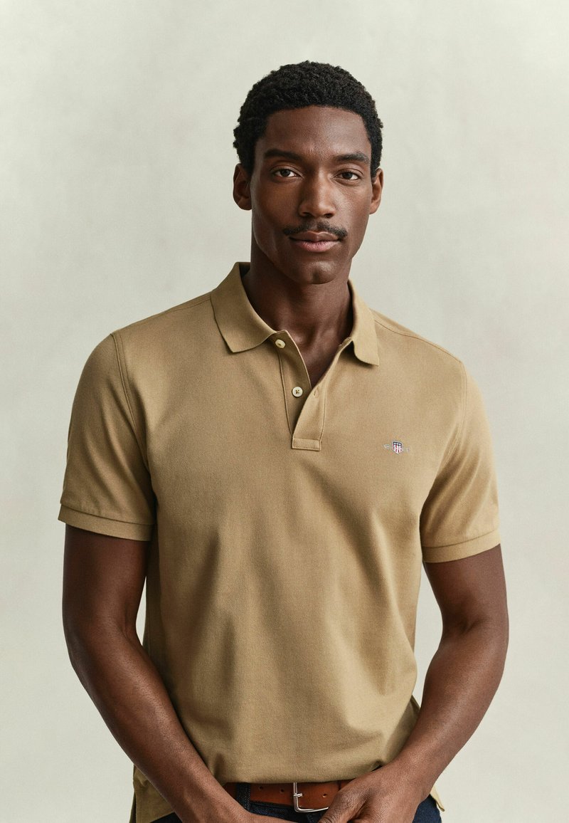 Jeune homme aux cheveux courts et bouclés avec une moustache, portant un polo beige, debout devant un fond clair uni.