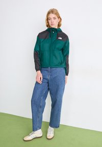 Tume rohelise värvi tuulejope mustade aktsentidega, millel on kapuuts ja elastsed mansetid, koos laia sinise teksaga ja beežide tossudega.