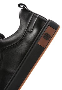 Zapatilla negra de cuero deslizante con textura suave, diseño esculpido y suela de goma marrón que presenta un acento de logo en el lateral.