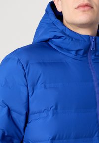 Chaqueta inflable azul con capucha, con una textura suave y un sutil logo de Calvin Klein. Cierre de cremallera y diseño acolchado horizontal.