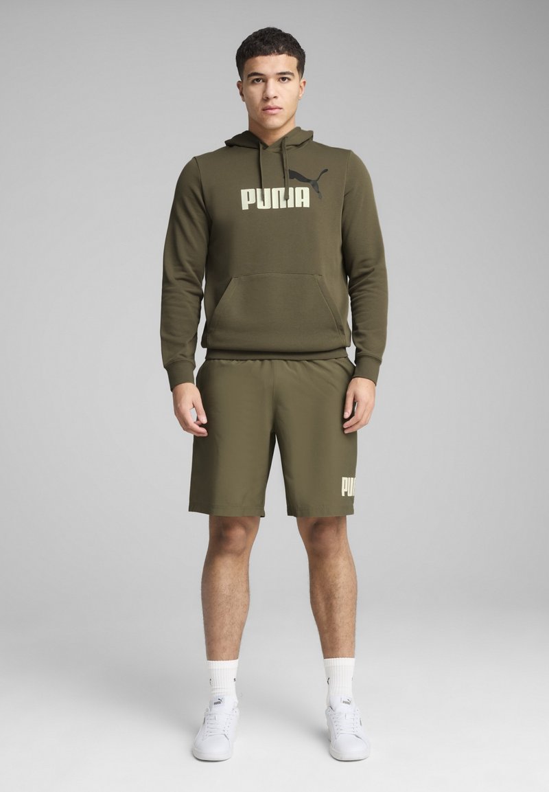 Olivgrünes Set aus Kapuzenpullover und Shorts aus weichem Material, mit einer Fronttasche und einem cremefarbenen Logo auf der Brust und den Shorts. Weiße Sneakers.